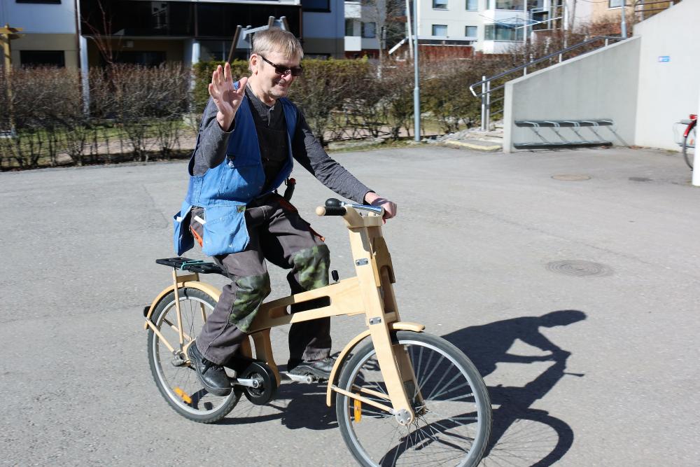Pekka Jääskeläinen ajaa puurakenteiseilla polkupyörällä ja heilauttaa kättään samalla tervehdykseksi.